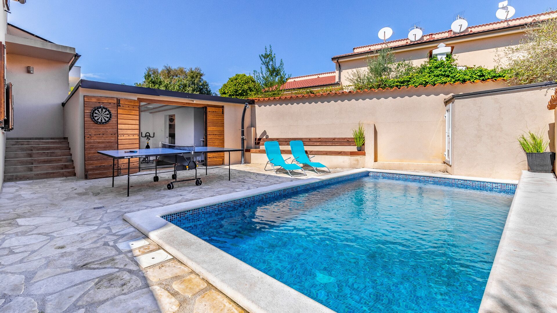 A swimming pool with a stone deck, a ping pong table, and two blue lounge chairs.