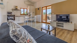 A modern living room with a sofa, a TV, a dining area with chairs, and a kitchen with cabinets and a window.