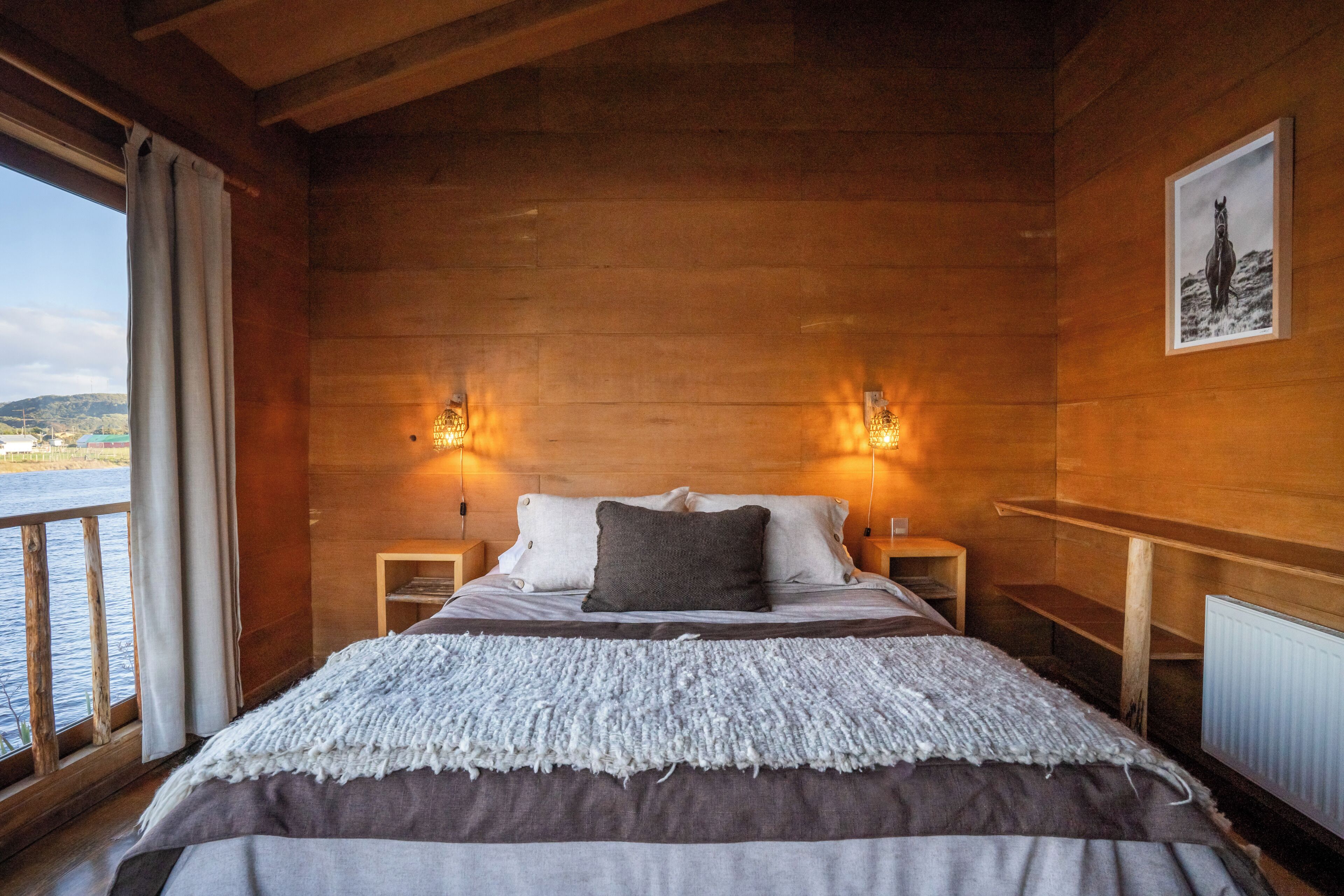 Un dormitorio de cabaña de madera con una cama, dos mesitas de noche, una mesilla auxiliar, un cuadro en la pared y vista al agua.