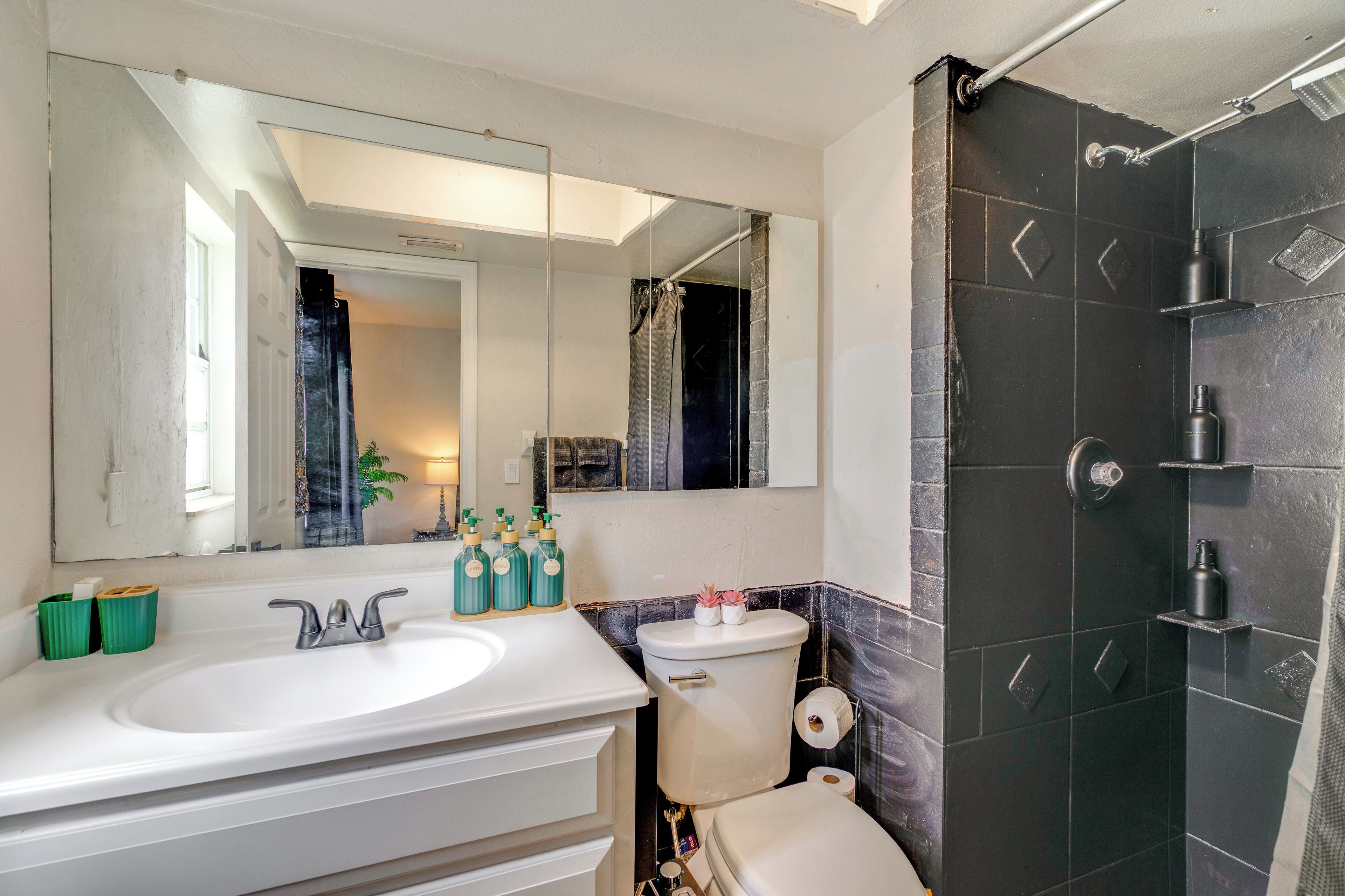 Une salle de bain avec un lavabo blanc, un miroir et une douche dont le sol est recouvert de carreaux noirs.