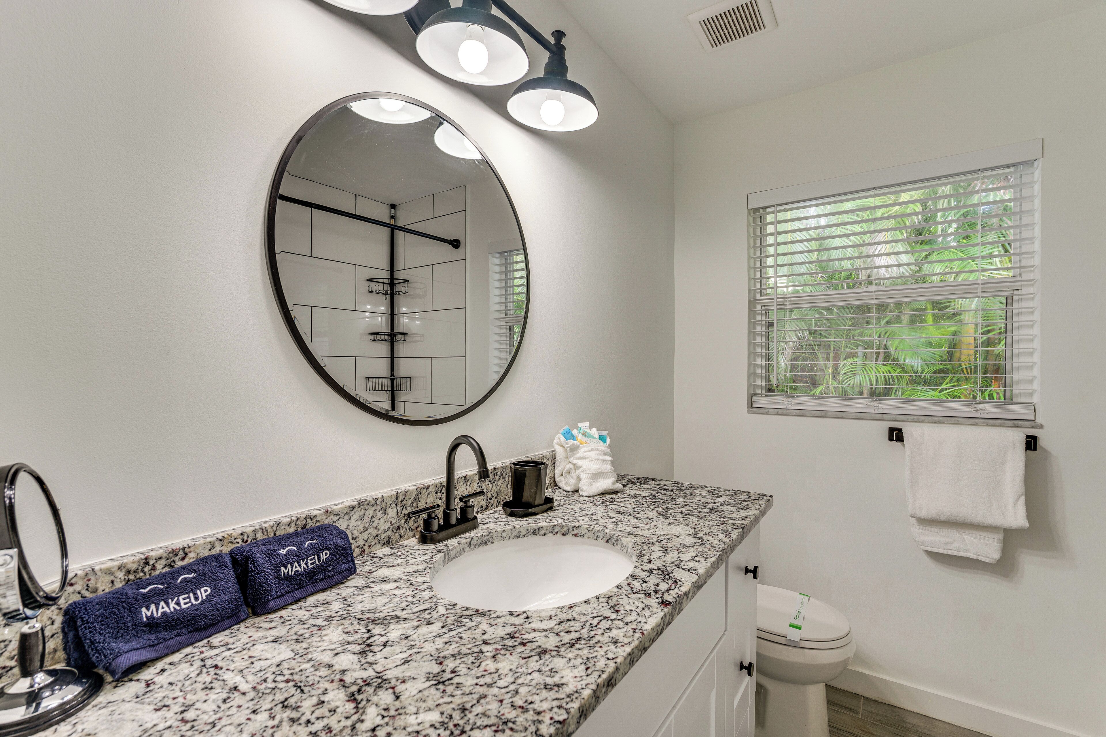 A bathroom with a granite countertop, a round mirror, a sink, and a window with blinds.