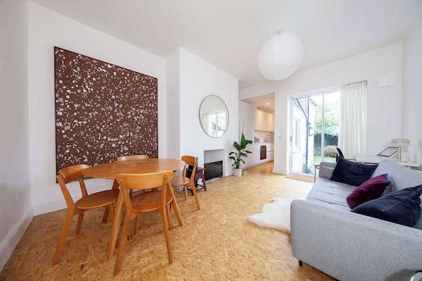 A modern living room with a wooden dining table and chairs, a grey sofa with cushions, a large abstract painting, and a fireplace.