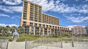Ein mehrstöckiges Hotel mit flachem Dach und Balkonen, umgeben von einem Sandstrand und Palmen.