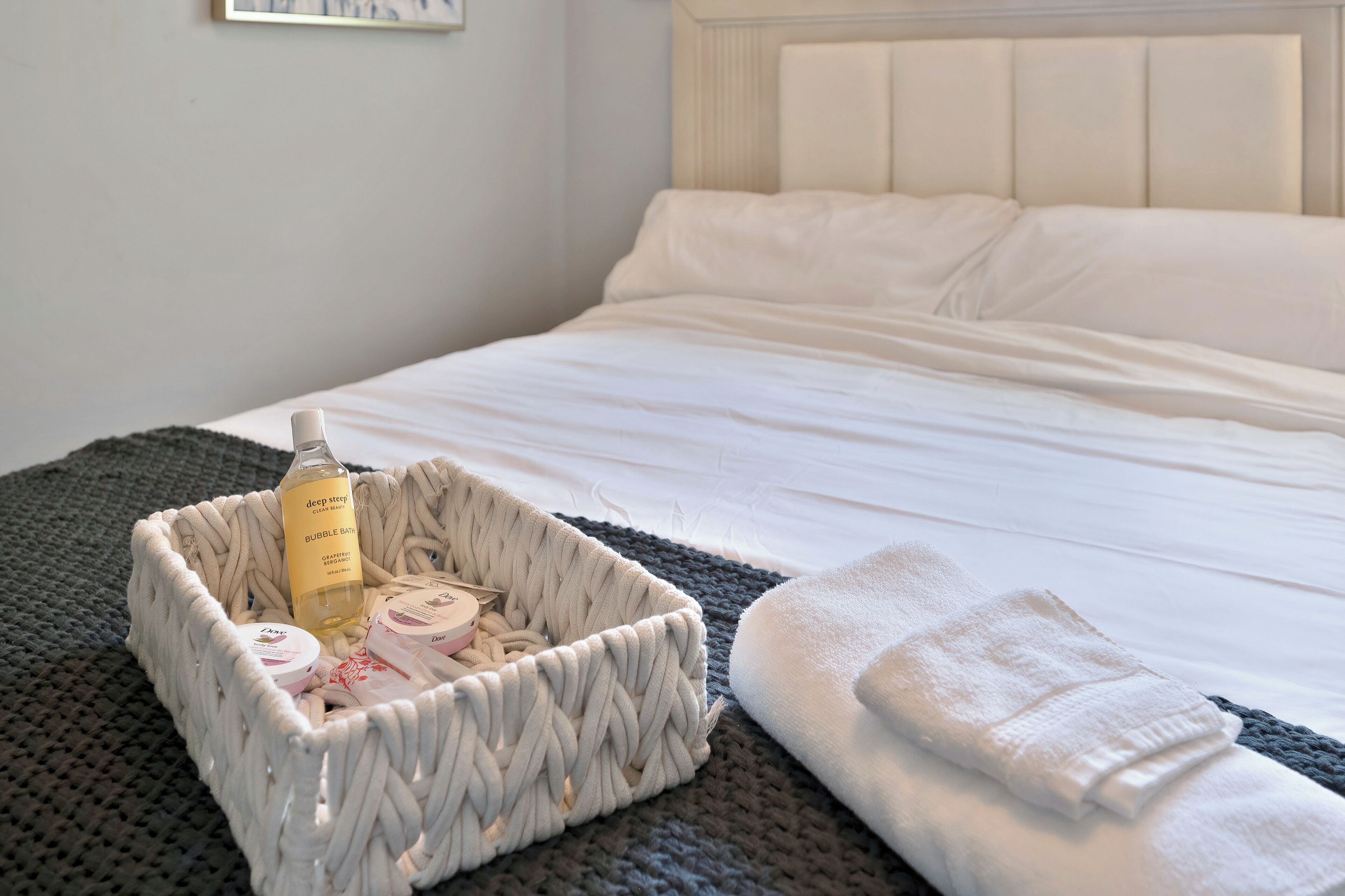A neatly made bed with white linens, a woven basket with toiletries, and a folded towel on a dark gray bedspread.