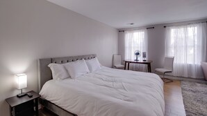 Une chambre bien rangée, avec un grand lit, une table de chevet avec une lampe, une petite table avec un ventilateur et deux chaises.