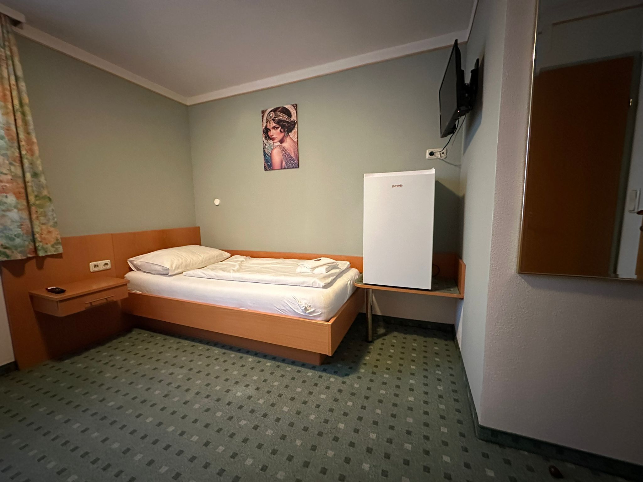 A small, single-bed room with a wooden headboard, a bedside table, a wall-mounted TV, and a refrigerator.