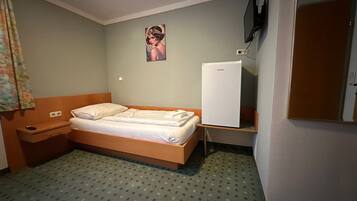 A small, single-bed room with a wooden headboard, a bedside table, a wall-mounted TV, and a refrigerator.