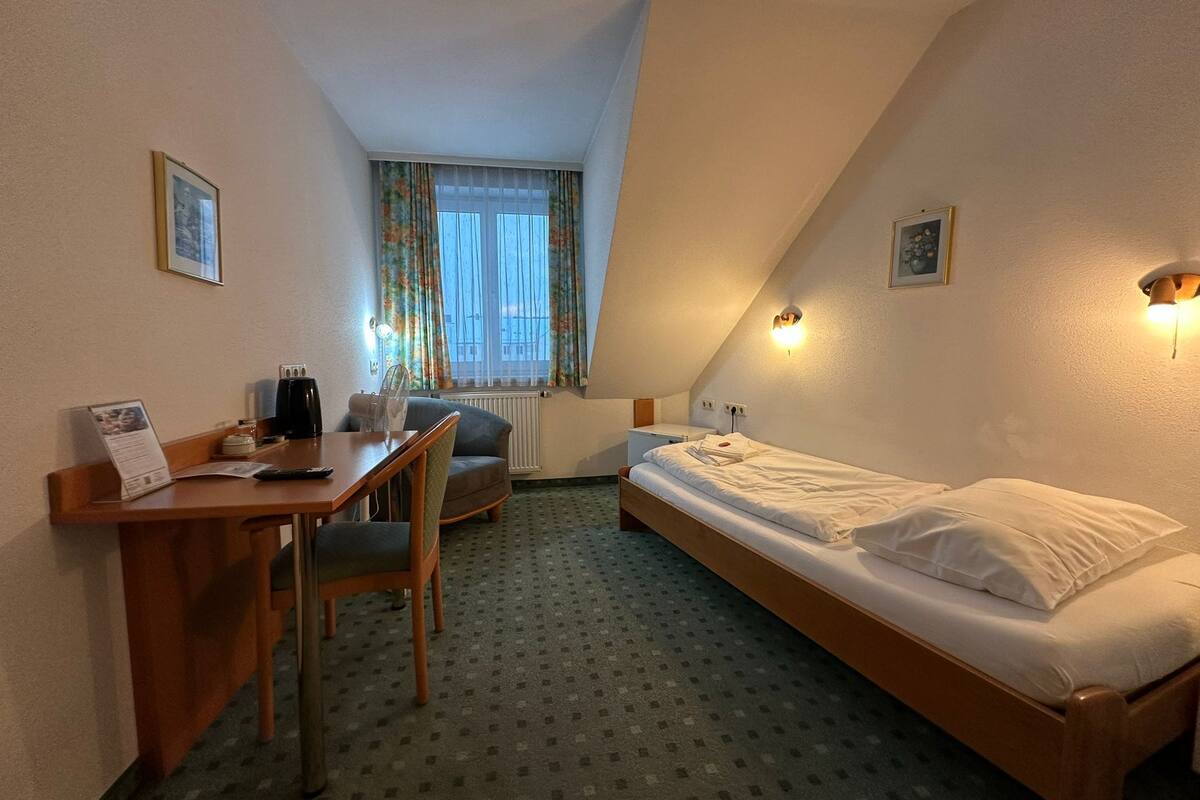Ein kleiner, gut beleuchteter Raum mit einem Bett, einem Schreibtisch, einem Stuhl und einem fenster mit Vorhängen.