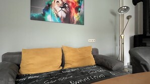 Una habitación con un sofá gris, dos cojines color mostaza y una manta negra con el texto 'Sweet Home'. En la pared, un cuadro de un león colorido.