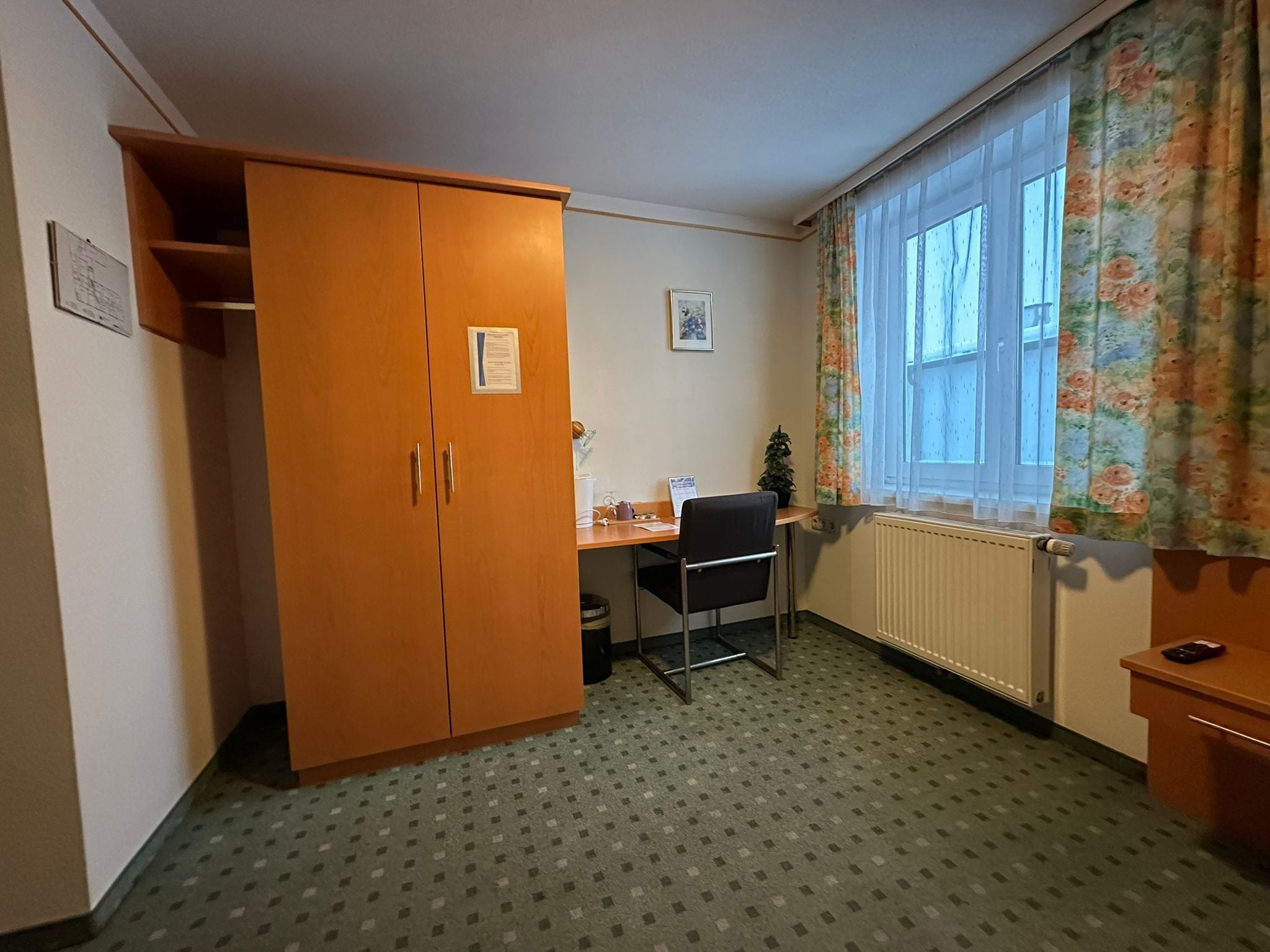 Ett litet rum med en trägarderob, ett skrivbord med en stol, ett element och ett fönster med blommiga gardiner.