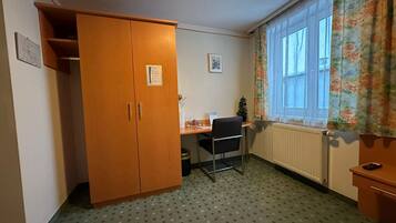 Ett litet rum med en trägarderob, ett skrivbord med en stol, ett element och ett fönster med blommiga gardiner.
