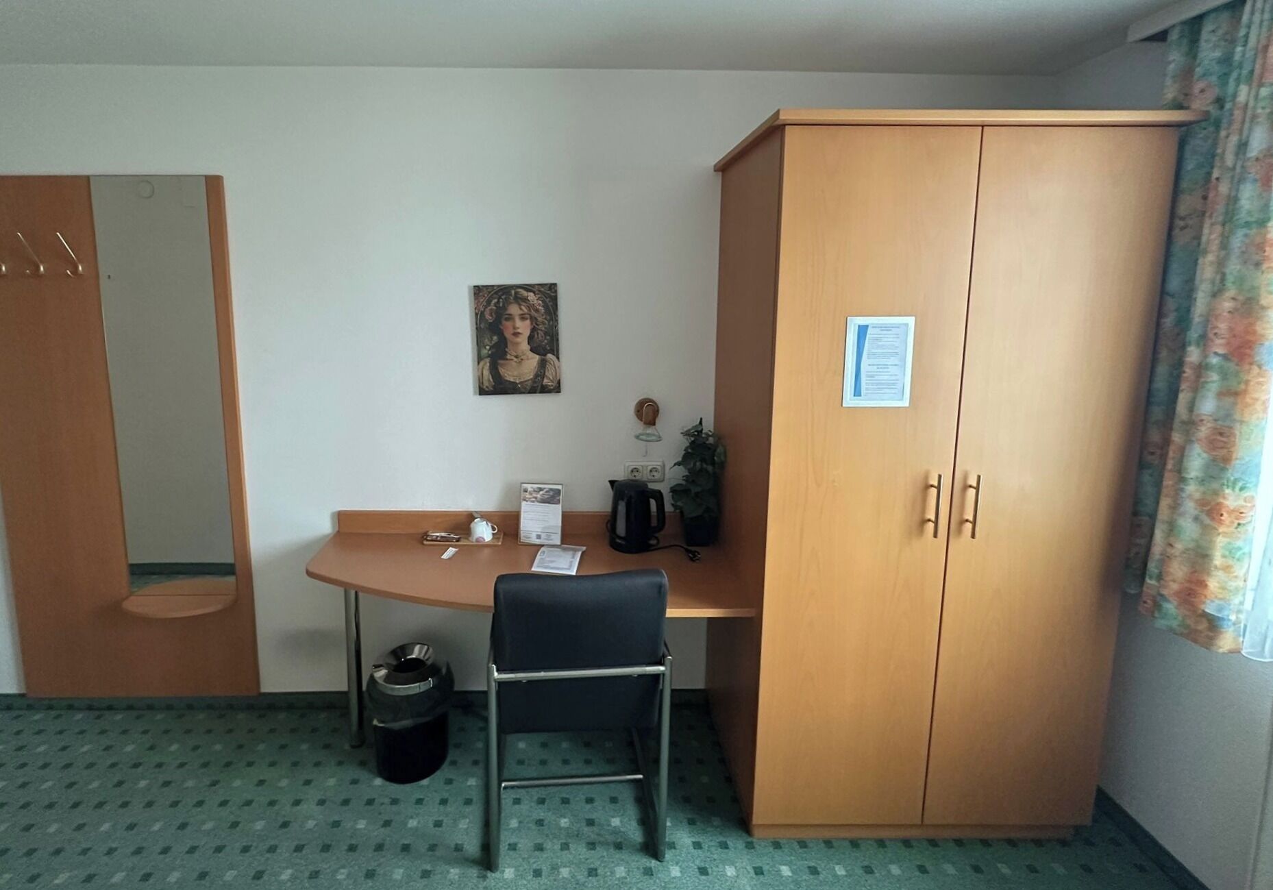 Une chambre d’hôtel avec une armoire en bois, un bureau avec une chaise, une petite poubelle, un tableau encadré au mur et une fenêtre avec des rideaux.