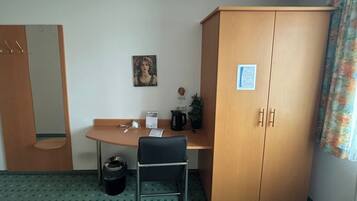 Une chambre d’hôtel avec une armoire en bois, un bureau avec une chaise, une petite poubelle, un tableau encadré au mur et une fenêtre avec des rideaux.