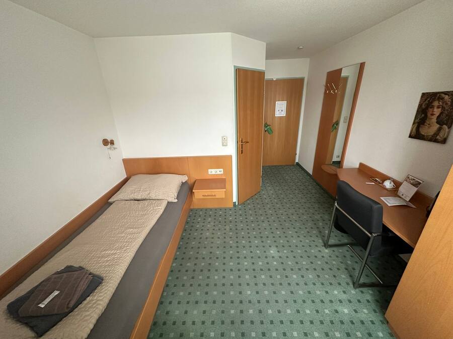 A hotel room with a single bed, a desk, a chair, a mirror, and a door leading to another room.