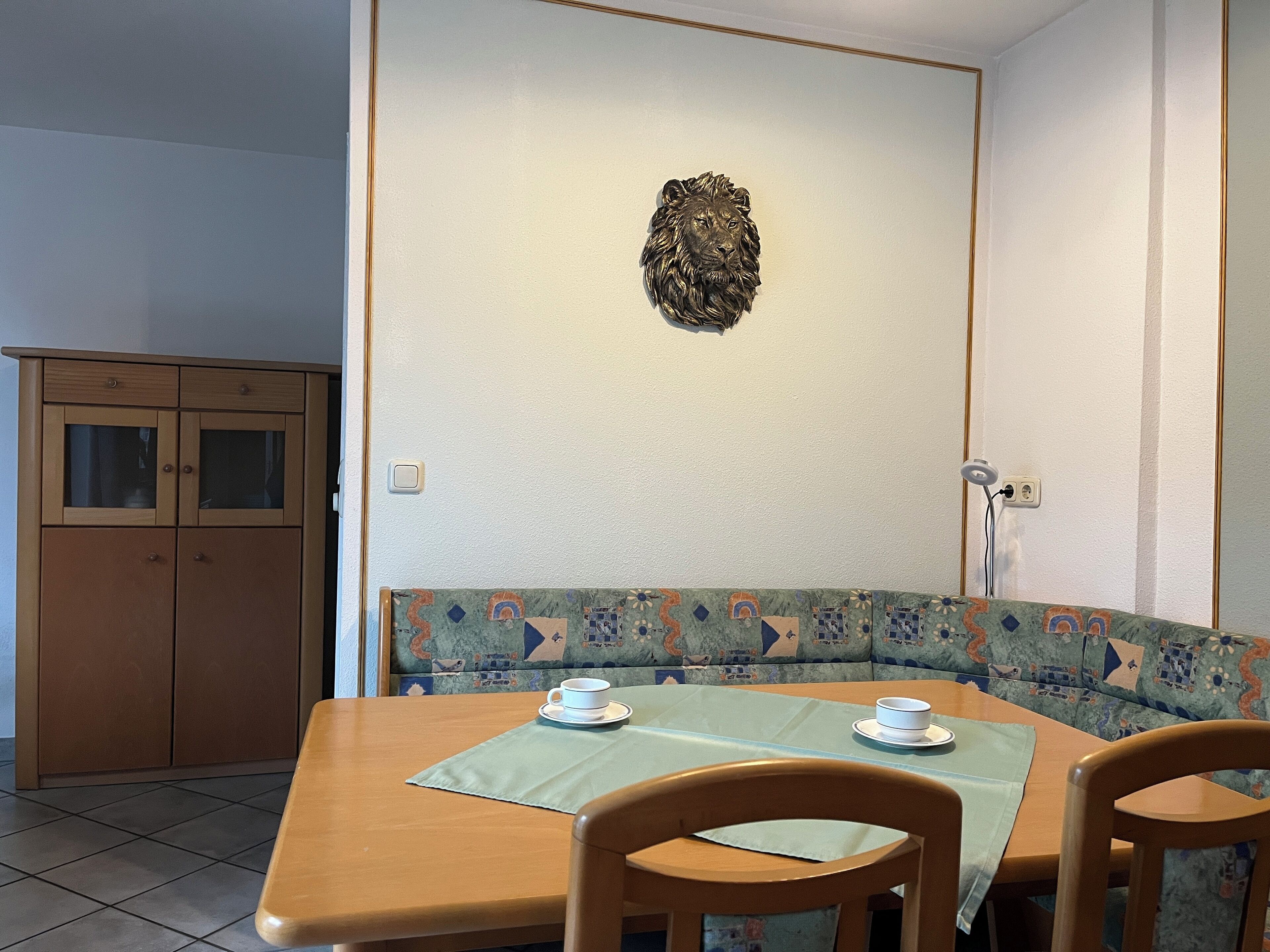 Un comedor con una mesa de madera, dos sillas, un banco con cojín estampado y una cabeza de león decorativa en la pared.