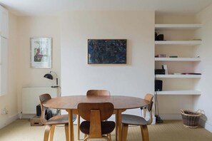 A dining area with a round wooden table and chairs, a radiator, a framed picture, and a shelf with books and a basket.