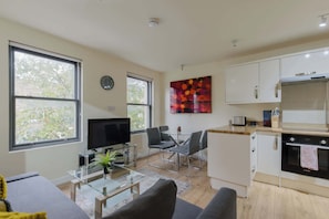 Ein modernes Wohnzimmer mit Flachbildfernseher, Glastisch und Sofa. Es gibt eine Essecke mit Glastisch und Stühlen sowie eine Küche mit weißen Schränken und einem Bosch-Ofen.