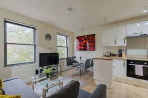 A modern living room with a flat-screen TV, a glass coffee table, and a sofa. There is a dining area with a glass table and chairs, and a kitchen with white cabinets and a Bosch oven.