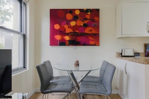 Une cuisine moderne avec une table à manger au plateau en verre, deux chaises grises, un tableau abstrait aux couleurs vives et un four encastré.