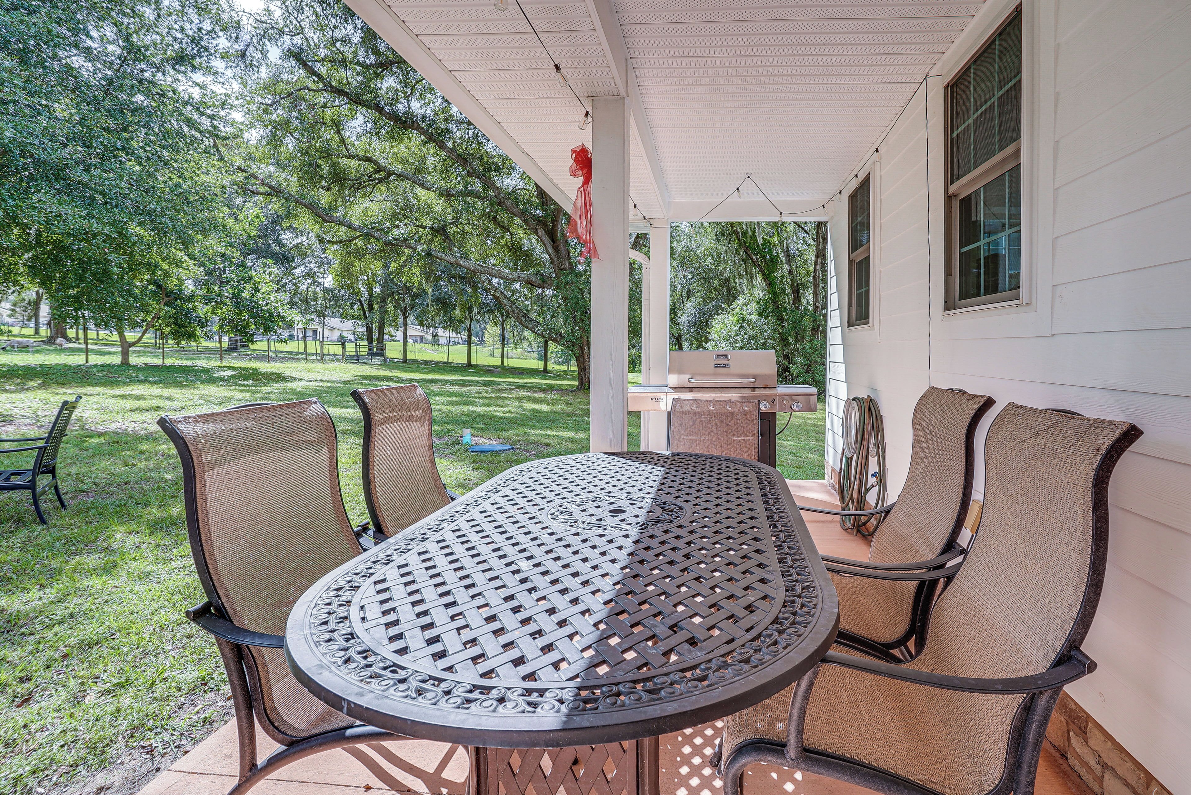 A covered patio with a round table and four chairs, a grill, and a view of a grassy area with trees.
