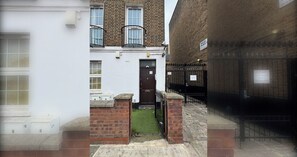 Une ruelle étroite, avec un bâtiment blanc sur la gauche, un bâtiment en briques au centre et un portail en métal noir sur la droite.