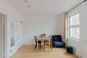 Une pièce comprenant une table à manger en bois, deux chaises, un canapé bleu, un petit tabouret vert et une fenêtre avec des rideaux.