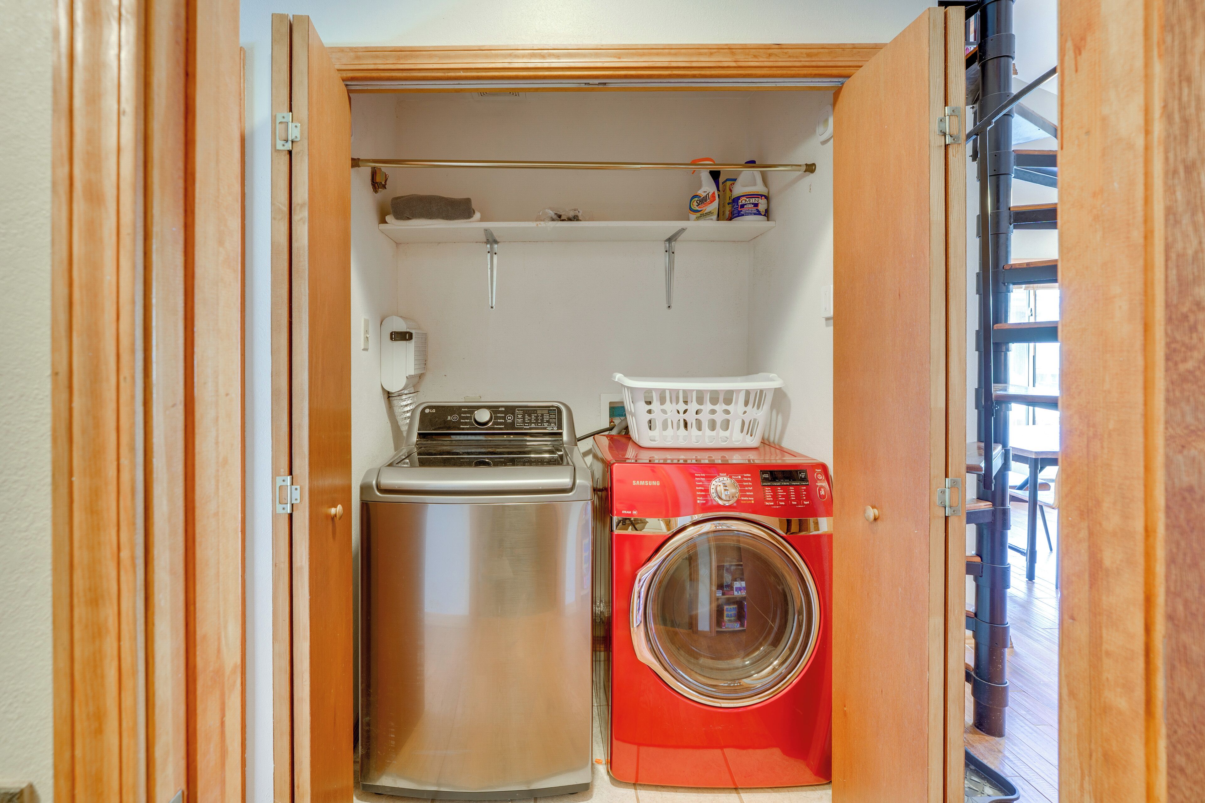 Une petite buanderie équipée d’un lave-linge et d’un sèche-linge en acier inoxydable, d’un panier à linge blanc et d’une étagère contenant des produits de nettoyage.
