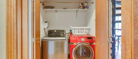 A compact laundry room with a stainless steel washer and dryer, a white laundry basket, and a shelf with cleaning supplies.
