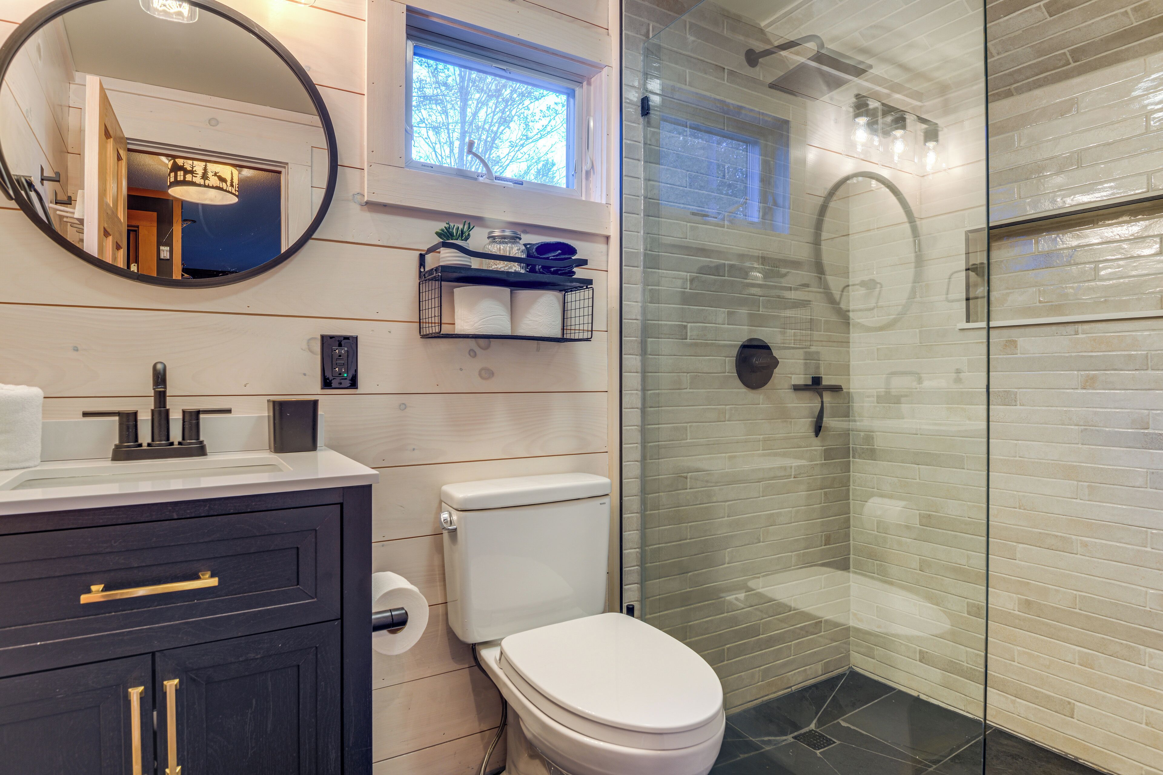 A bathroom with a white toilet, a dark vanity with gold handles, a round mirror, and a glass-enclosed shower area with a tiled wall.