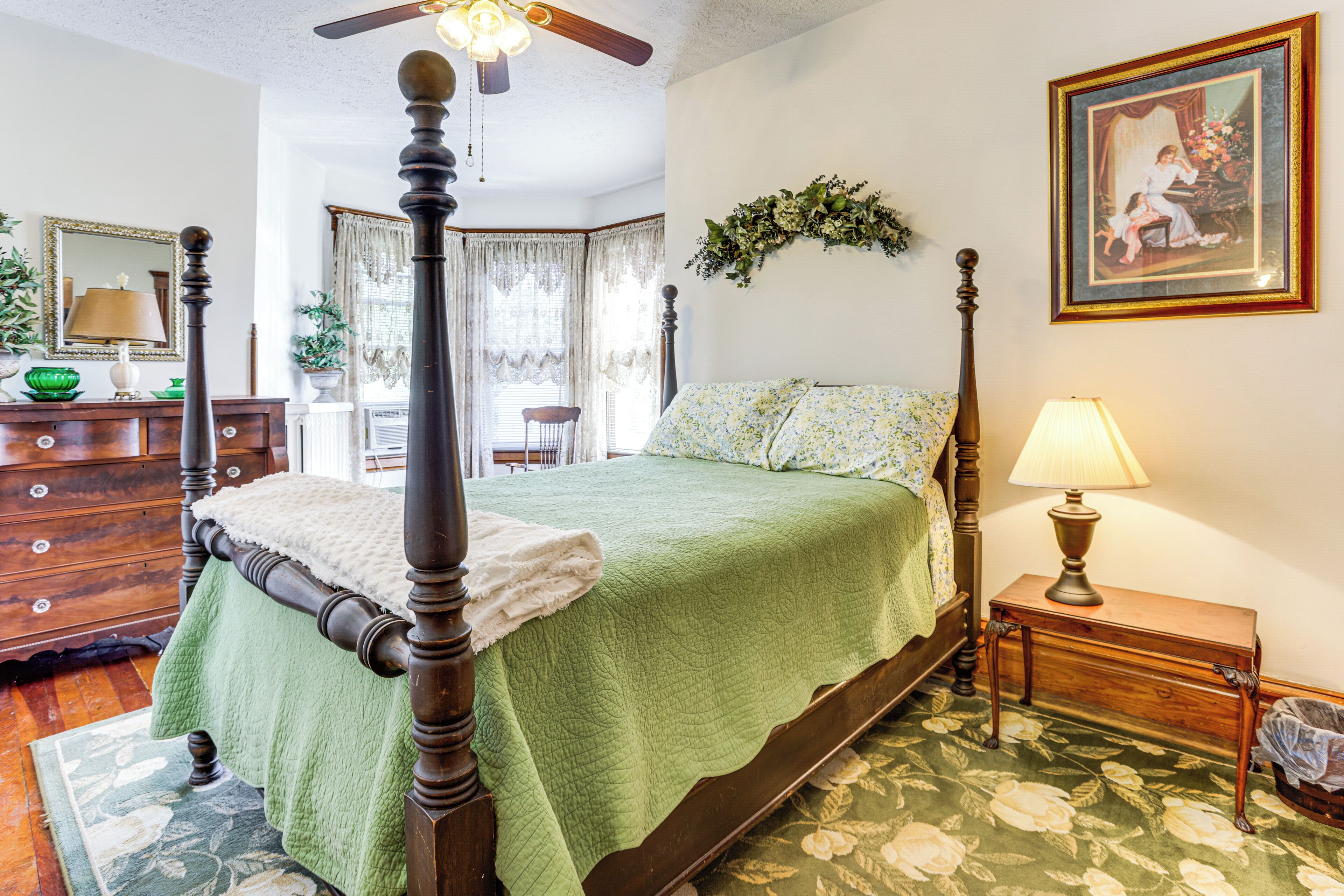 A four-poster bed with a green bedspread, a wooden nightstand with a lamp, a dresser with a green bowl, and a floral-patterned rug.