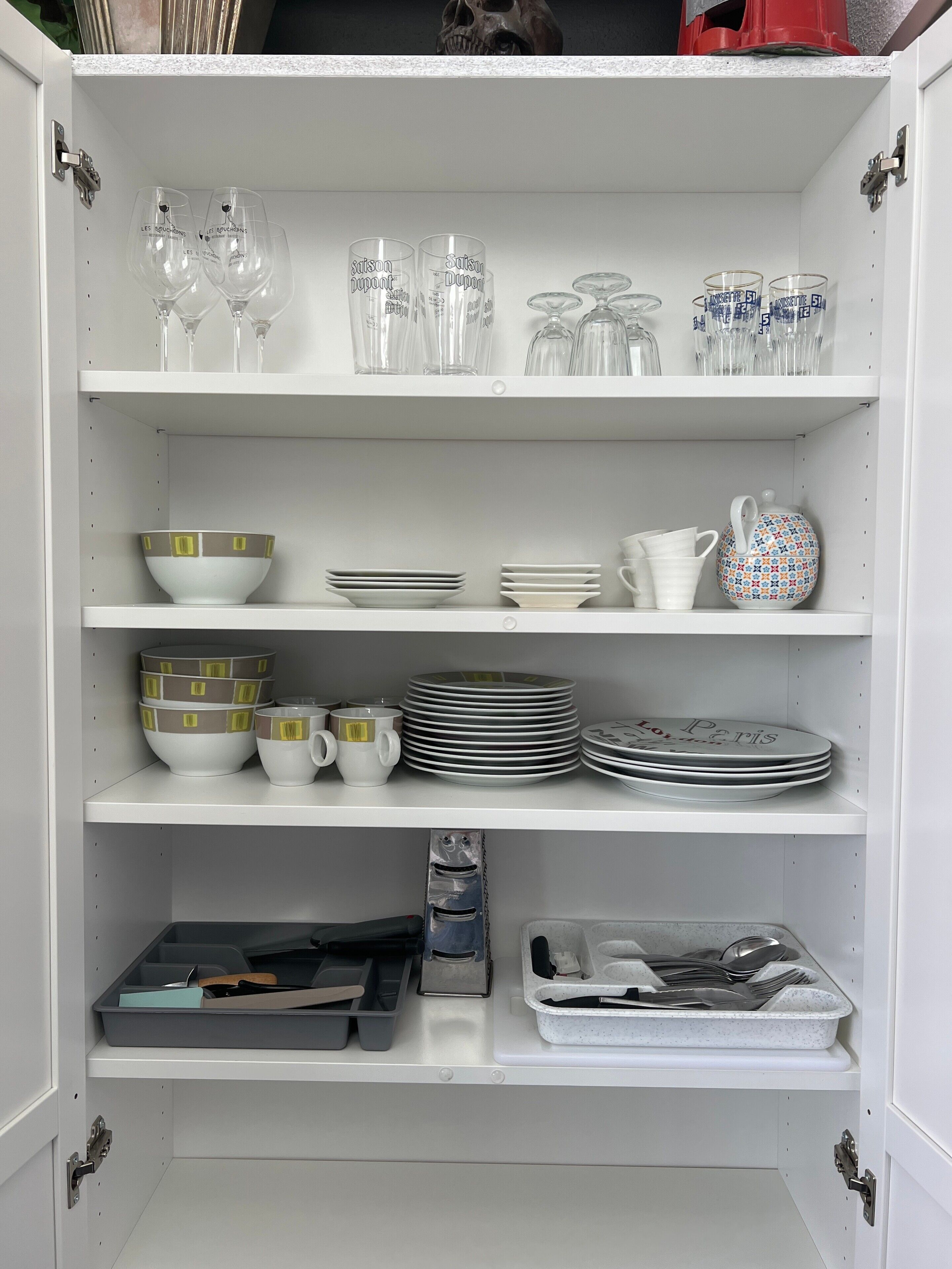A white cabinet with shelves containing various kitchenware, including wine glasses, bowls, plates, and utensils.