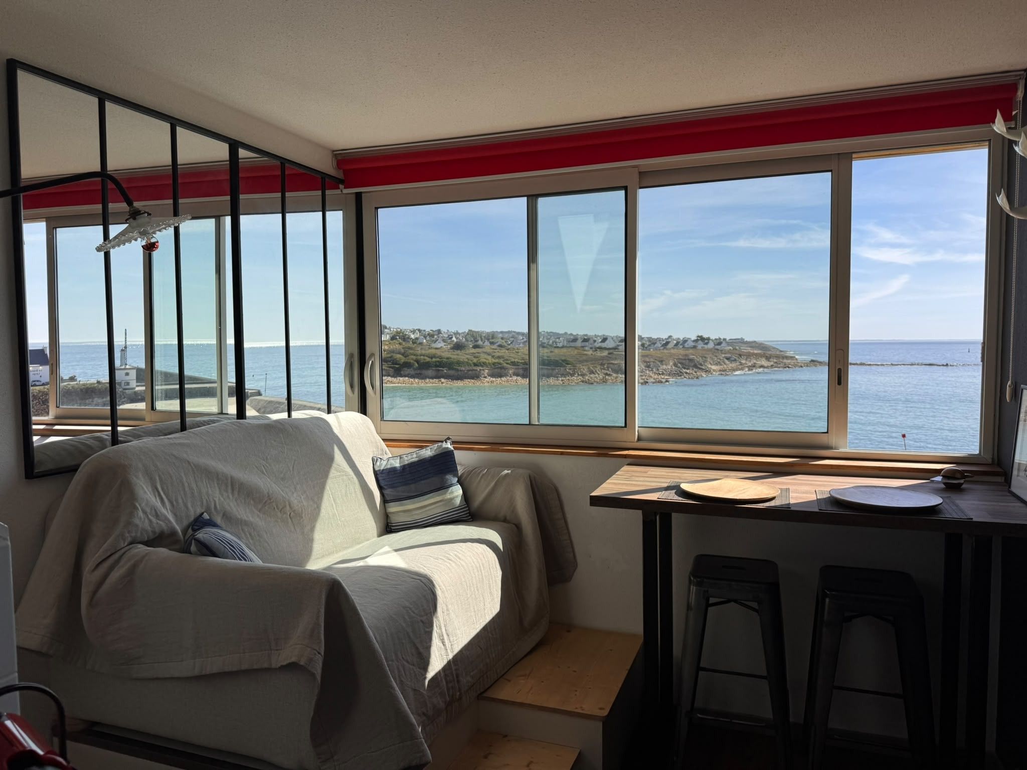 A room with a sofa, a dining table with chairs, and large windows offering a view of the sea and a distant shoreline.