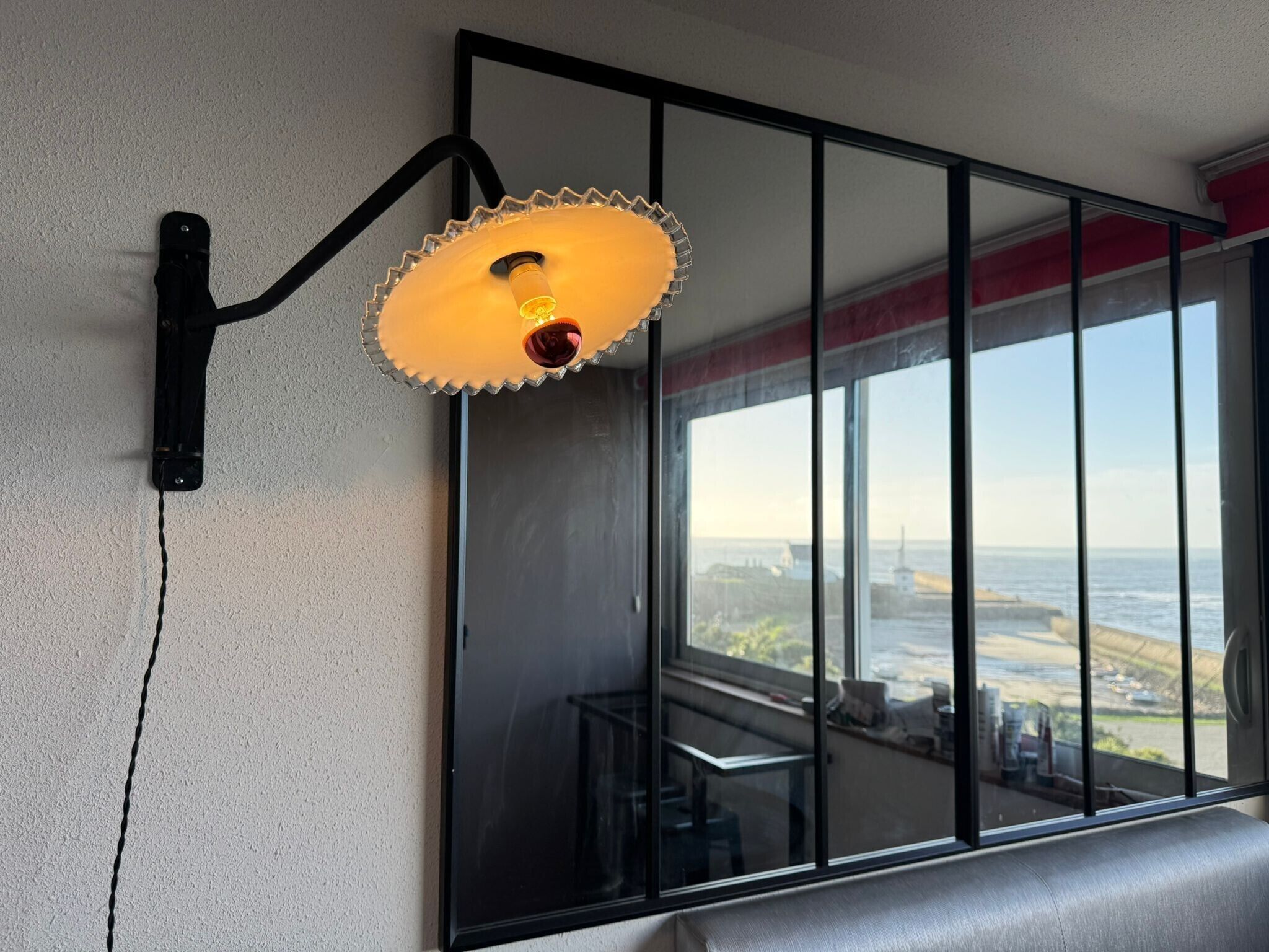 A wall-mounted light fixture with a unique design, a large window with a view of the sea, and a reflection of the room's interior.