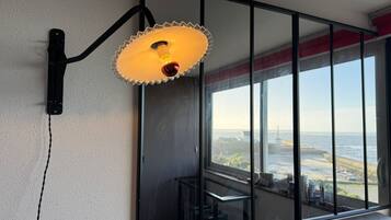 A wall-mounted light fixture with a unique design, a large window with a view of the sea, and a reflection of the room's interior.