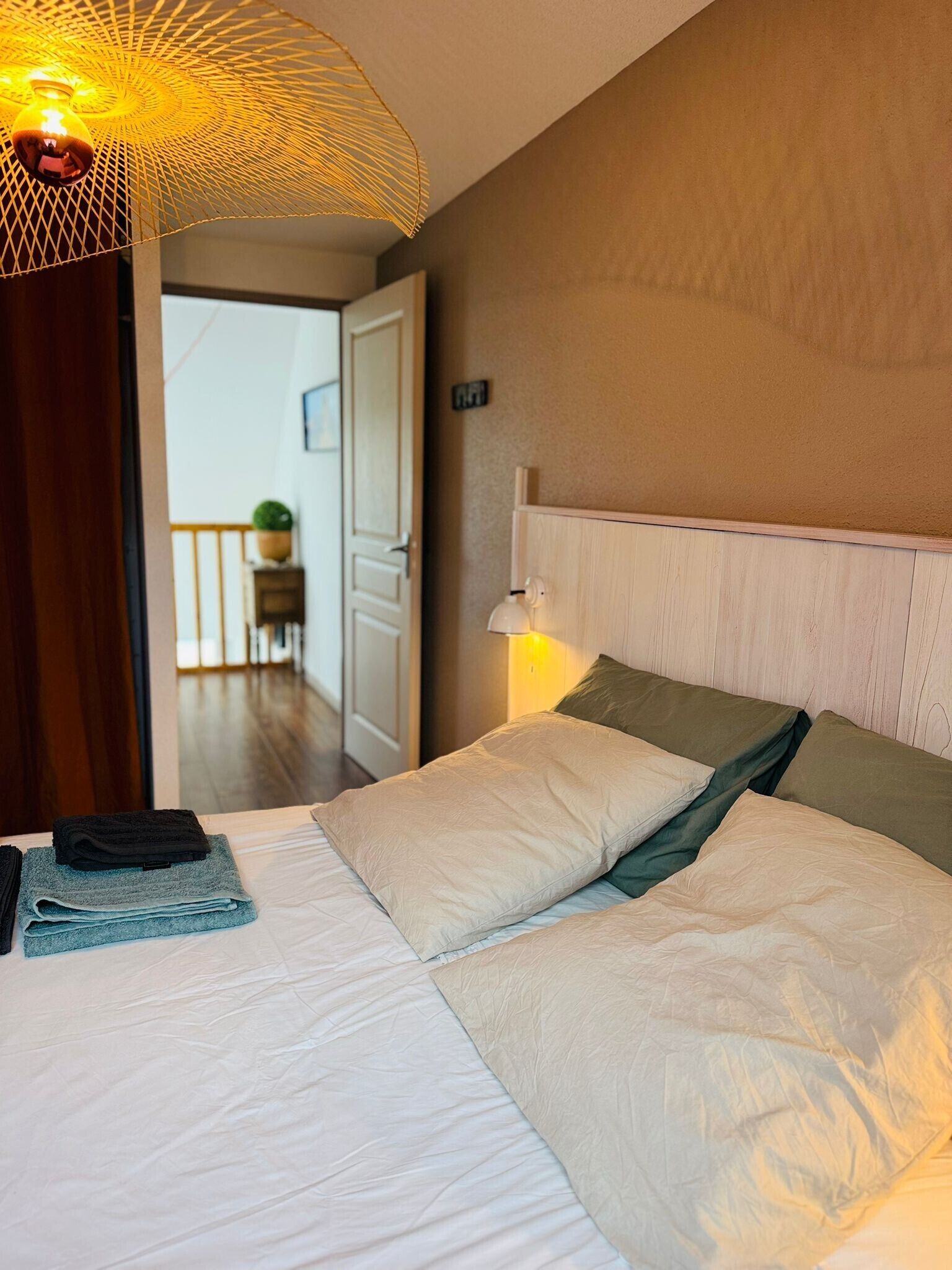A bedroom with a bed, pillows, a bedside lamp, a hanging light fixture, and a view of a hallway.