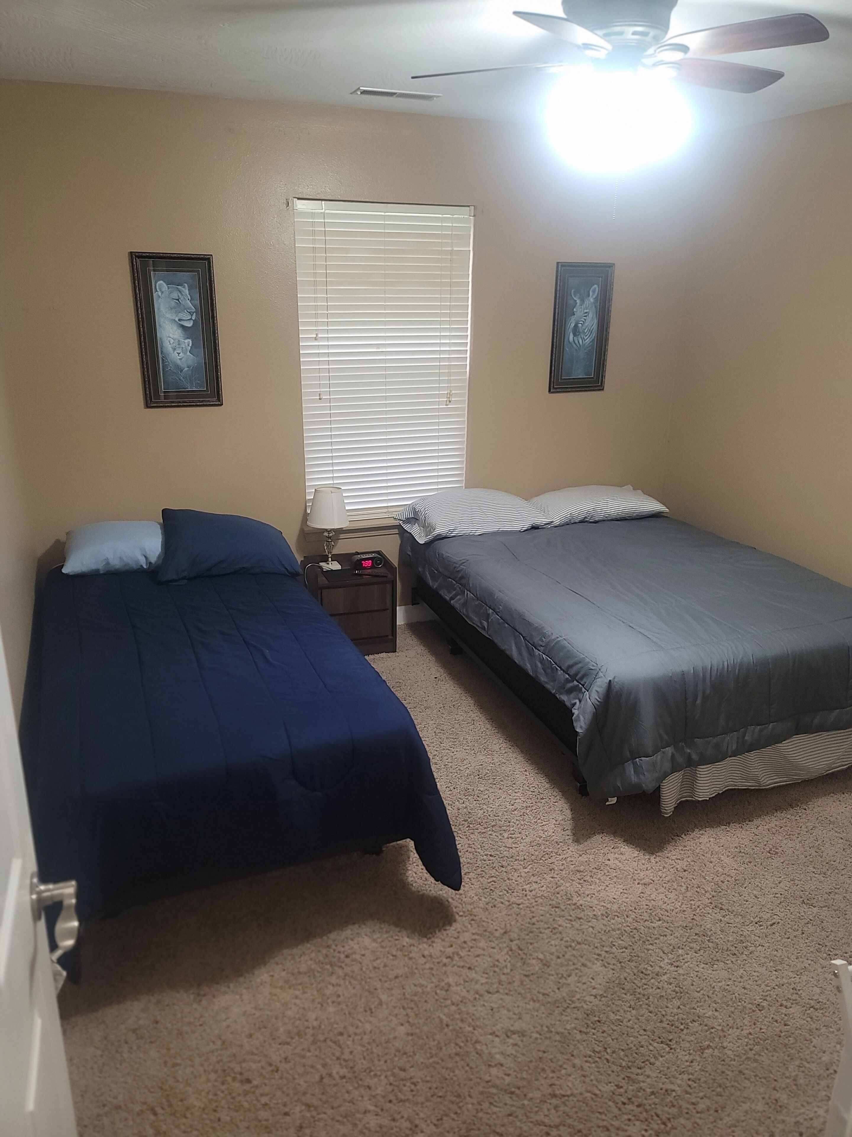 Une chambre avec deux lits, un ventilateur de plafond, une table de chevet avec une lampe et une fenêtre avec des stores.