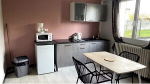 A compact kitchen with a microwave, coffee maker, and a small refrigerator. There is a dining table with chairs and a window with curtains. - Apartment T1 Paisible (Limoges)