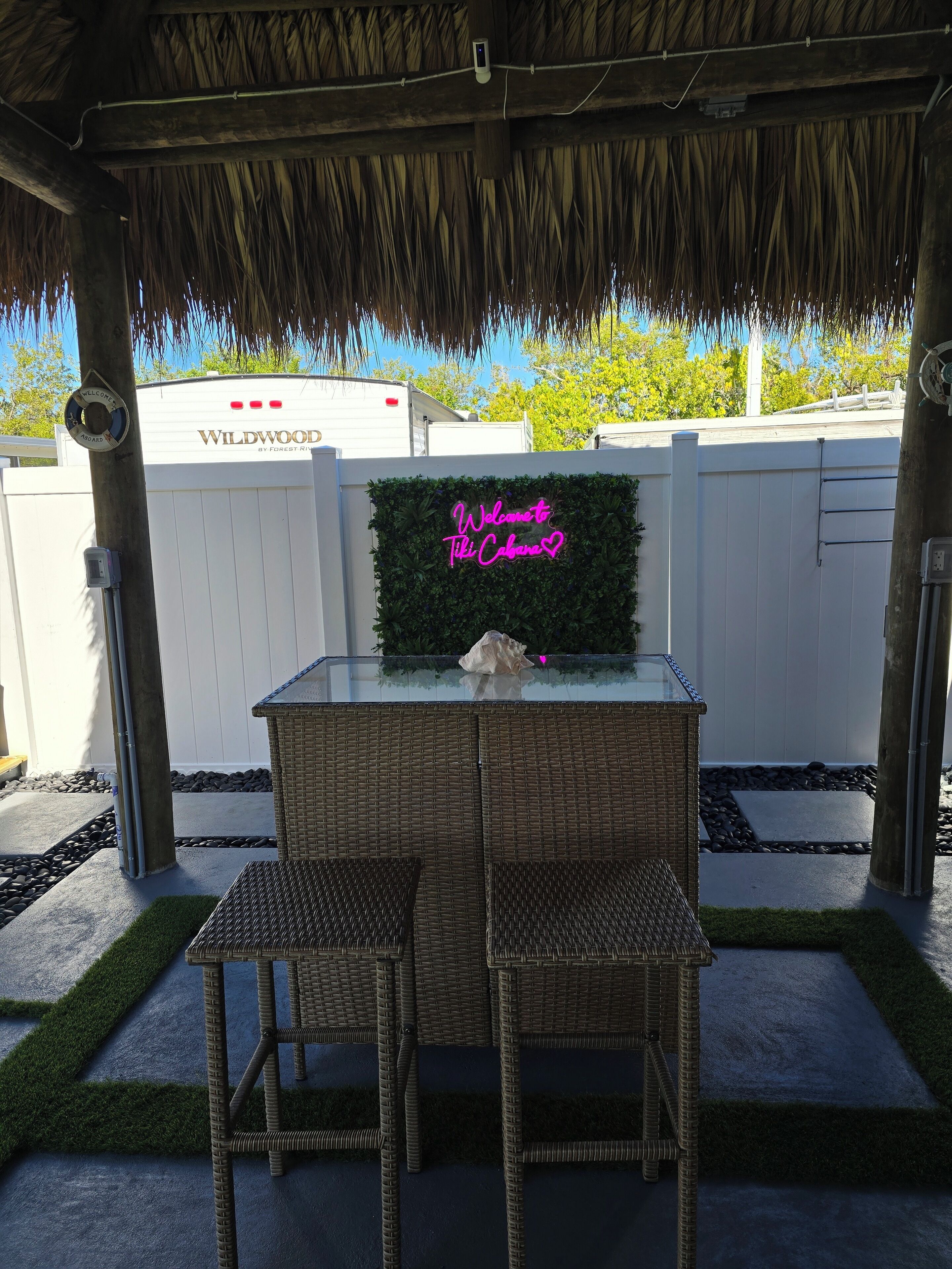 An outdoor seating area with wicker chairs and a glass table, a neon sign reading "Welcome to Tiki Cabana", and a white fence in the background.