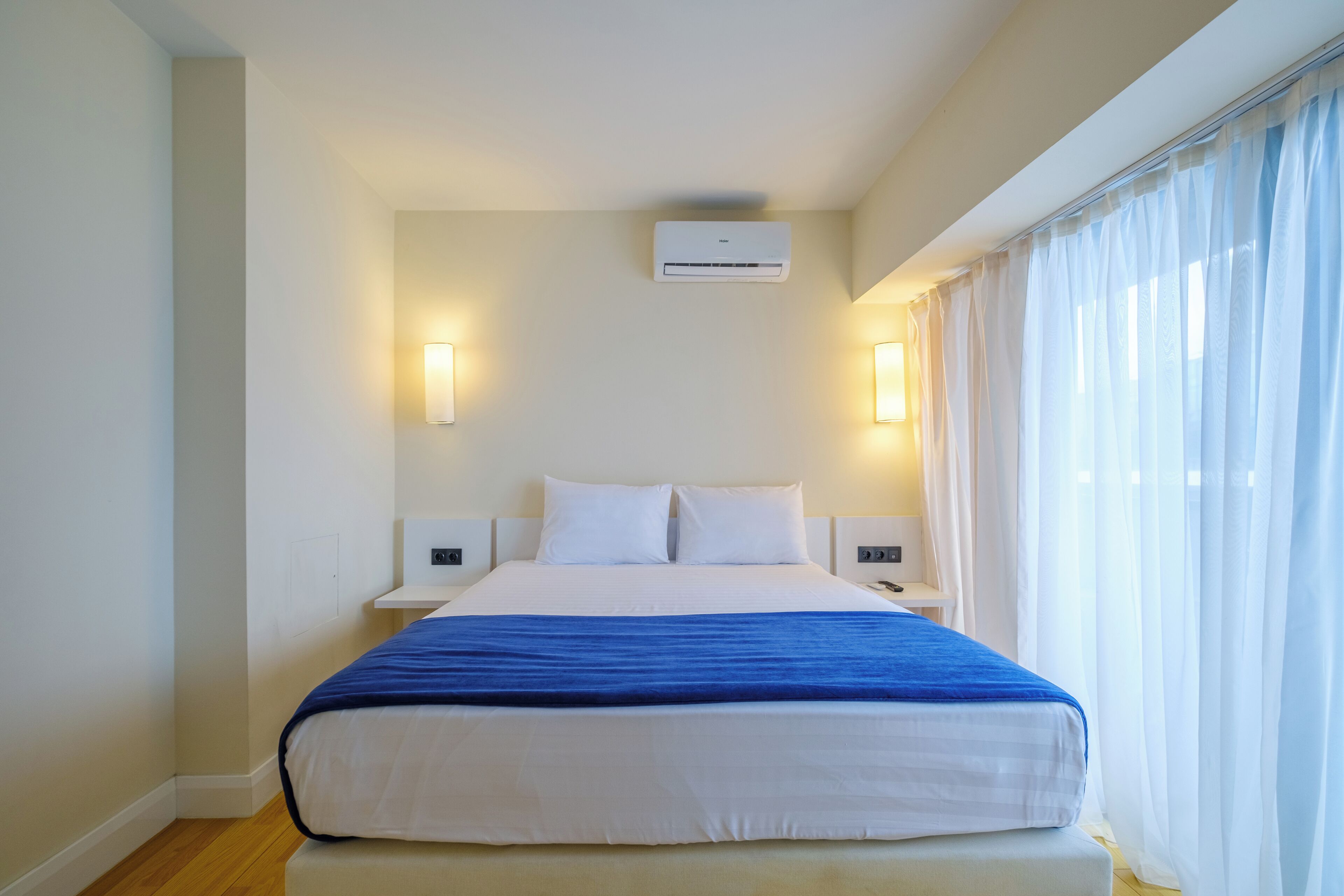 Una habitación de hotel con una cama grande, paredes blancas, un aire acondicionado, dos lámparas de noche y cortinas de gasa.