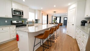A modern kitchen with white cabinets, a central island with bar stools, stainless steel appliances, and a fire extinguisher. - Walk to Stadium | Hot Tub • Fire Pit • Parking (Atlanta)