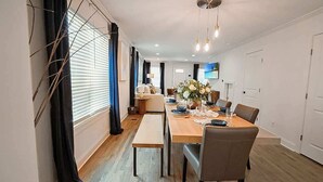 A modern dining area with a wooden table, chairs, and a bench. There is a TV mounted on the wall, and the room has a neutral color scheme. - Walk to Stadium | Hot Tub • Fire Pit • Parking (Atlanta)