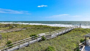 A coastal walkway with wooden railings leading to the beach, surrounded by grassy areas and a clear blue sky. - Legasea - Ocean Front home with direct Beach Access, Large Decks, great for larg (Carolina Beach)