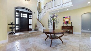 A spacious foyer with a grand staircase, a round wooden table with a vase, and a decorative wall piece. - Palm House GolfCourseViews Pool 5bd/5bth&Game Room (Naples)