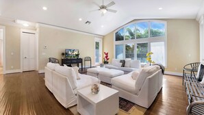 A spacious living room with a large window, a ceiling fan, and a flat-screen TV. - Palm House GolfCourseViews Pool 5bd/5bth&Game Room (Naples)