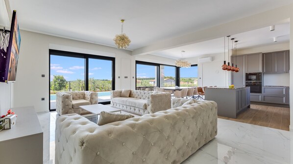 A modern living room with a large sofa, a dining area, and a kitchen with grey cabinets.
