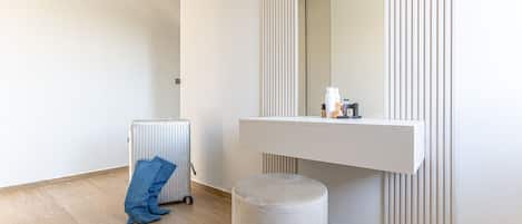 A hotel room with a radiator, a suitcase, a bench, and a vanity with toiletries.