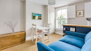 A living room with a blue sofa, a white dining table with chairs, a wooden cabinet, and a window with blinds. - Herne Hill Haven (London)