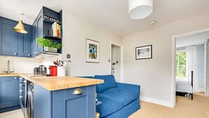 A modern kitchen with blue cabinets, a wooden countertop, and a built-in island. There is a blue sofa in the living area, a bookshelf with books, and a framed picture on the wall. - Herne Hill Haven (London)