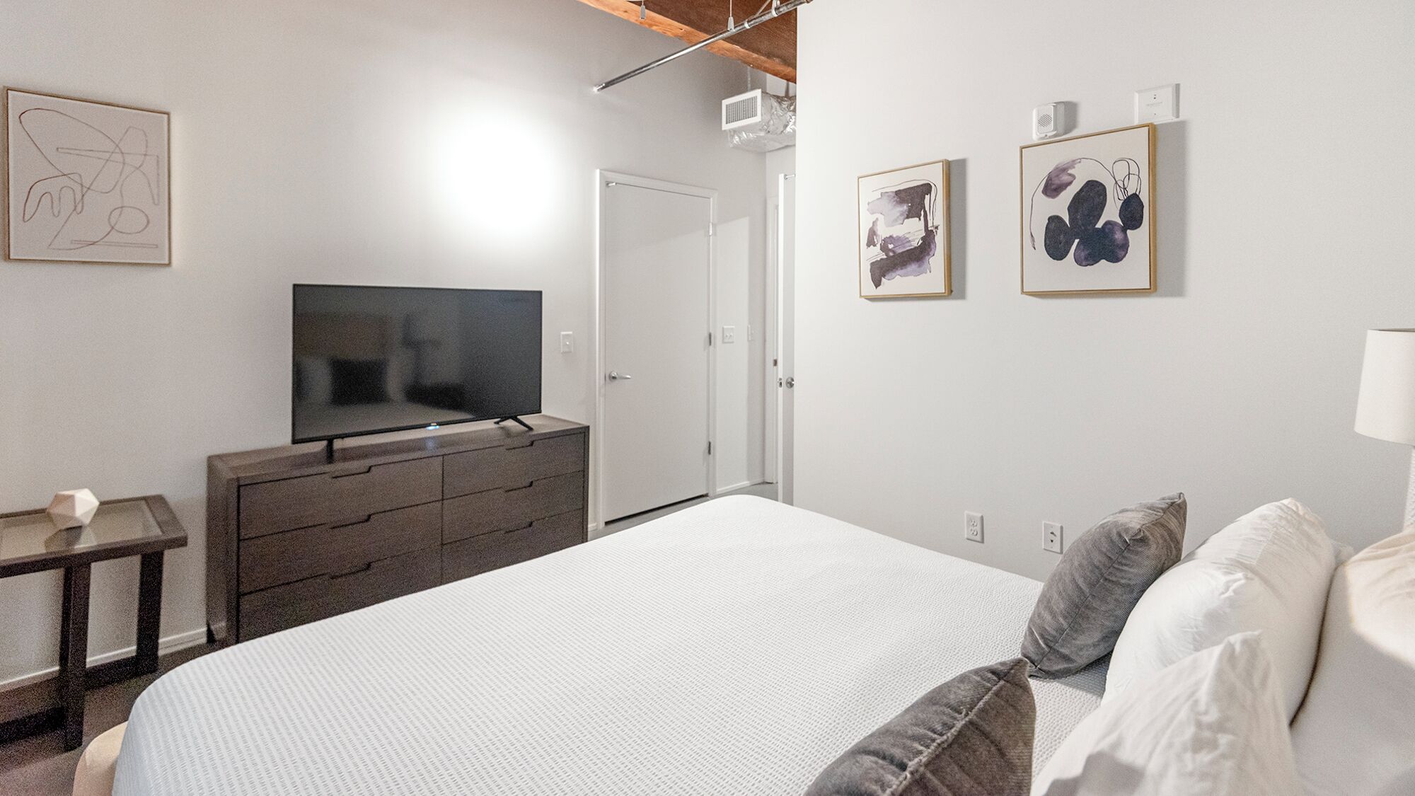 A hotel room with a bed, a TV on a dresser, a door, and two framed artworks on the wall.