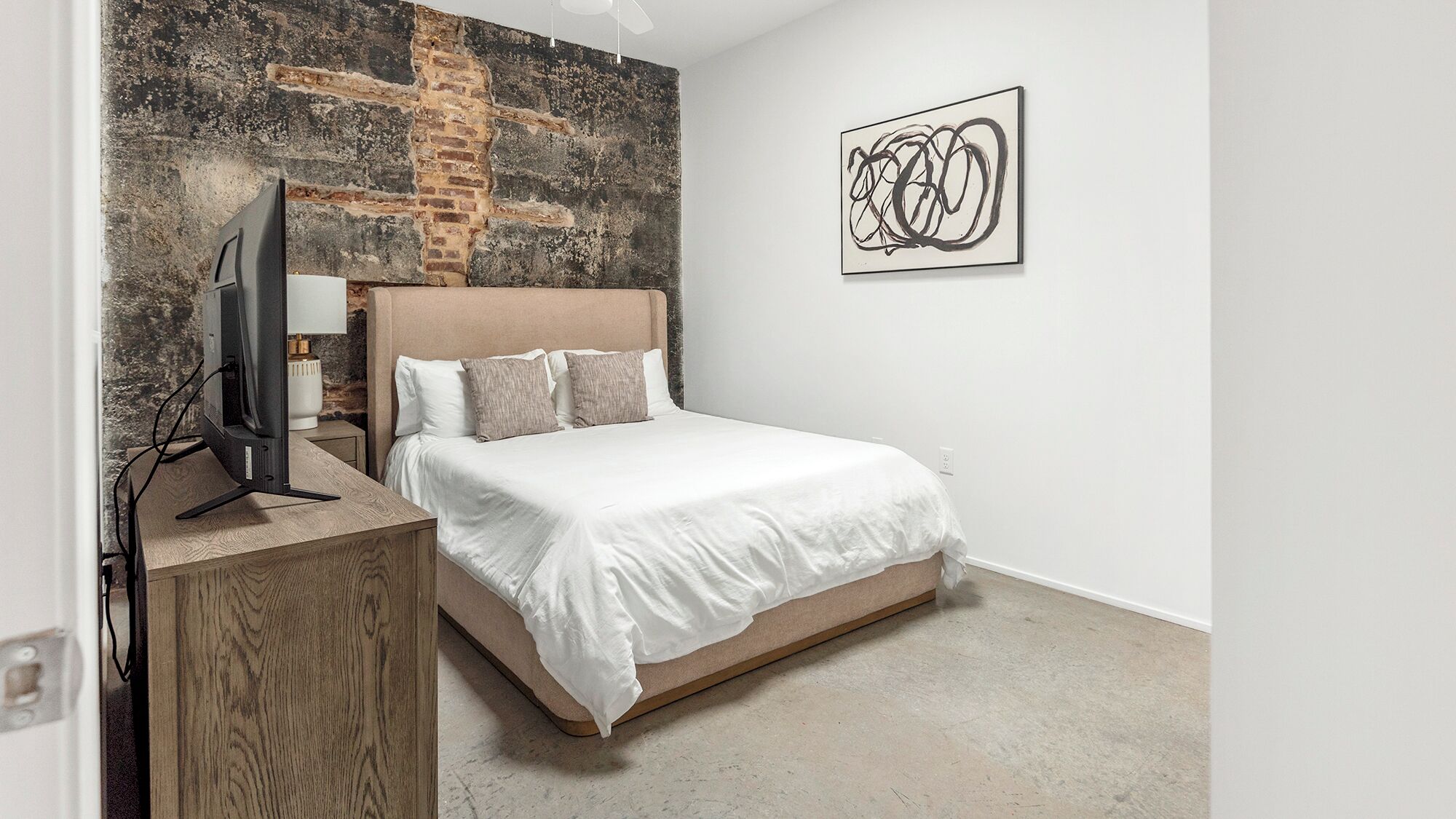 Un dormitorio con cama, televisor sobre un mueble de madera, pared de ladrillo y una obra de arte abstracta.